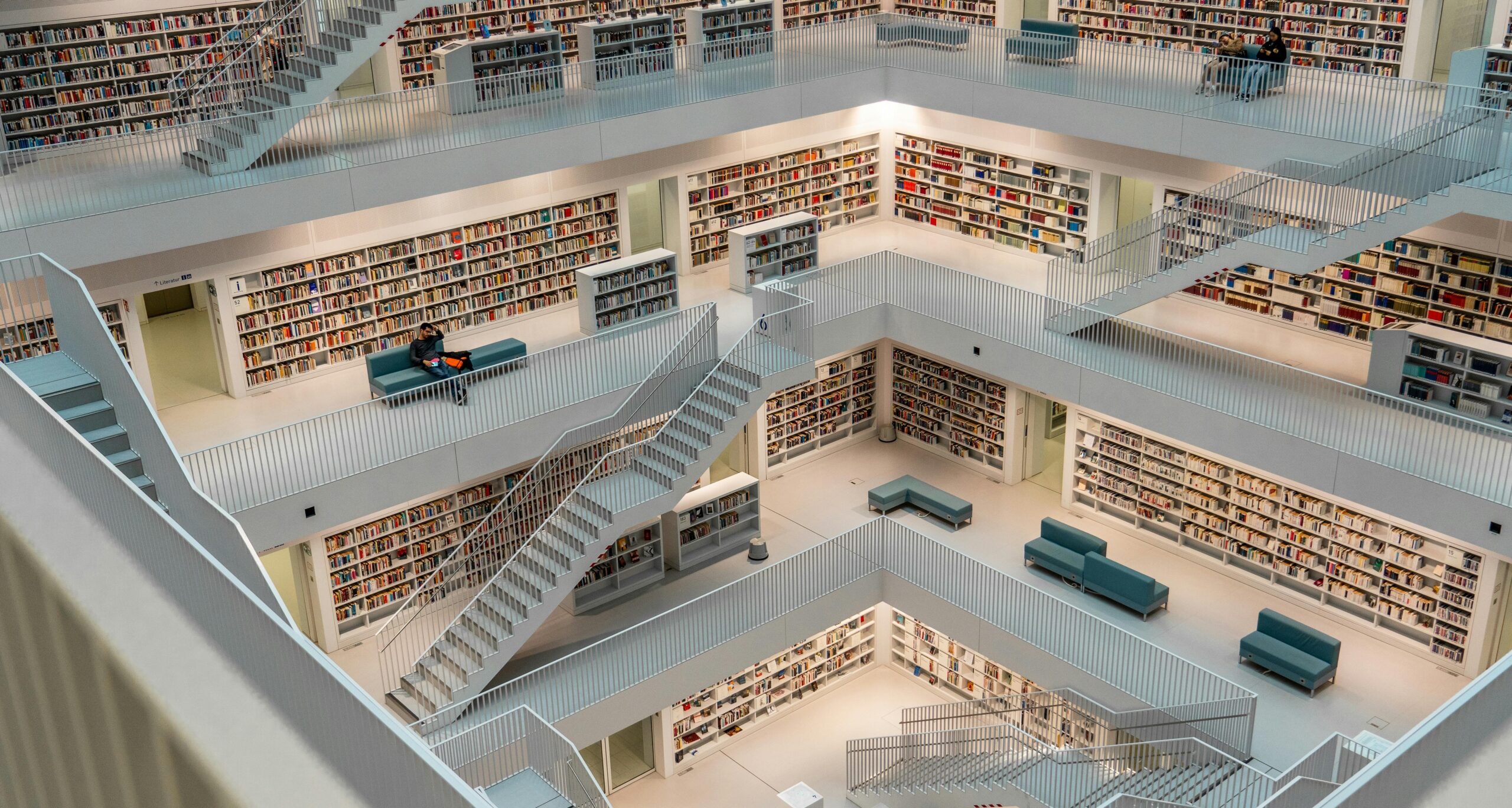 Modern library interior with multiple levels connected by white staircases. White walls lined with floor-to-ceiling bookshelves filled with books. Teal blue benches and couches scattered throughout. Minimalist design with white railings and bright lighting showing an open, airy architectural style